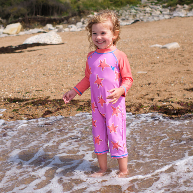 A young child smiles on a beach, paddling in the sea, wearing a UV Protective Surf Suit Pink Starfish.